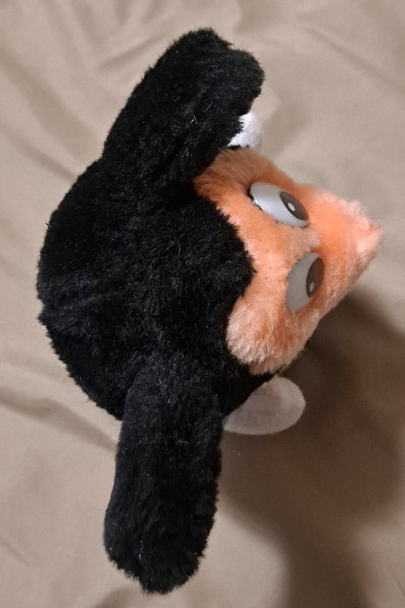 May include: A black and orange plush toy with large eyes. The toy has a black furry head and body with orange fur on the face and a white patch on the side.