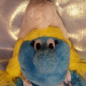 May include: A blue plush toy with a white hat and yellow hair. The toy has large, white eyes and a small, round nose.