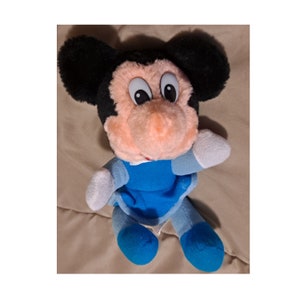 May include: A plush toy of Mickey Mouse in a blue dress. The toy has black ears, a pink nose, and large white eyes.