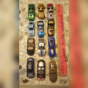May include: A collection of 12 toy cars, mostly blue and black, with some red, green, and gold. The cars are all different makes and models, and some have logos and writing on them. The cars are arranged in three rows of four, and are sitting on a wooden surface with a pink ruler.
