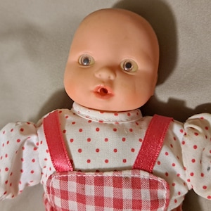 May include: A porcelain baby doll with a pink and white gingham dress and a white shirt with red polka dots. The doll has a pink blush and blue eyes.