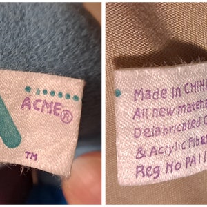 May include: A close-up of two white fabric tags with purple and green text. The first tag shows the logo for ACME with the text "ACME&reg; TM". The second tag reads "Made in CHINA All new materials Delabricated Cotton & Acrylic Fibers Reg No PA1163".