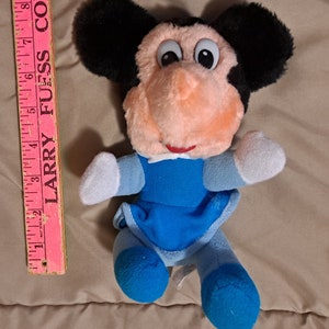 May include: A plush toy of Mickey Mouse wearing a blue dress with white trim. The toy has black ears, a pink nose, and large black eyes.