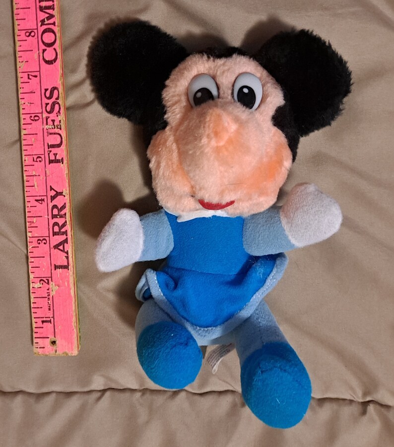 May include: A plush toy of Mickey Mouse wearing a blue dress with white trim. The toy has black ears, a pink nose, and large black eyes.