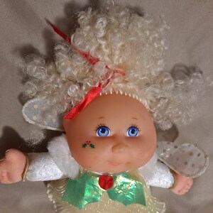 May include: A white doll with blonde curly hair wearing a white dress with a green and gold bow, white wings with gold polka dots, and a red ribbon in her hair. She has blue eyes and a holly design on her cheek.