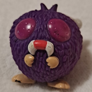 May include: A purple, round, cartoon-like figure with large, red eyes and a red mouth. It has small, tan arms and feet, and a spiky top. The figure has a fuzzy texture and a whimsical design, resembling a playful monster or creature.