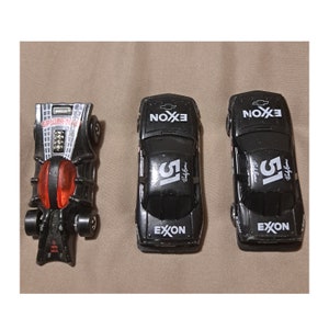 May include: Three toy cars are displayed. The first is a black Spider-Man car with red and silver accents. The other two are black race cars with the number 51, the Exxon logo, and the text "Randy Brown".