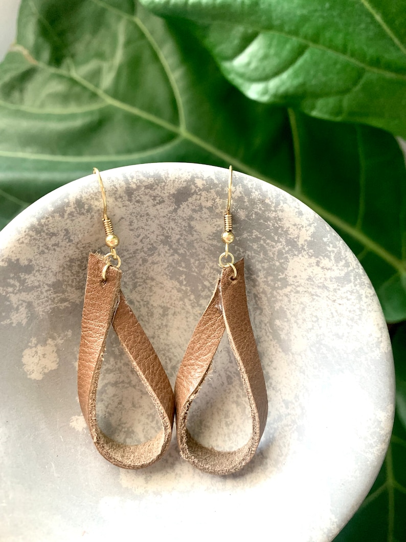 May include: A pair of tan leather teardrop earrings with gold-coloured ear wires. The earrings are looped into a minimalist design. The earrings are displayed on a light grey, textured surface. The earrings are a stylish accessory.
