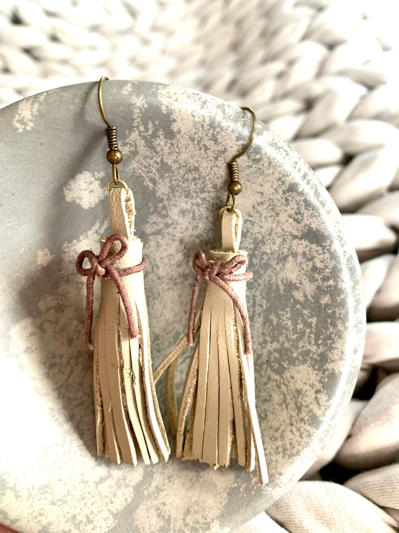 May include: A pair of cream-colored leather tassel earrings with a light pink bow detail and bronze hooks.