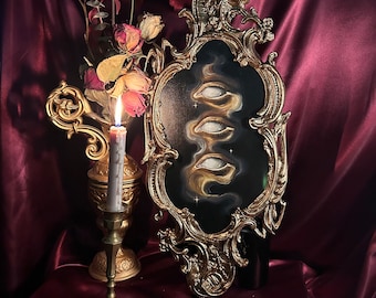 May include: A gold ornate frame with a black background and a painting of four white eyes with gold accents. The frame is on a dark red surface with a lit candle in a gold candlestick.