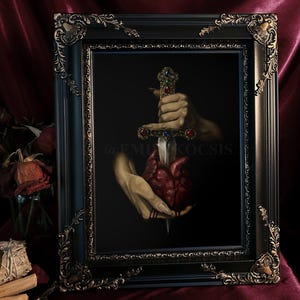 May include: A framed artwork depicts a hand holding a jeweled dagger piercing a heart. The heart is red, and the dagger has colorful gemstones. The frame is black with ornate gold detailing. The background is dark, with a hint of red fabric.