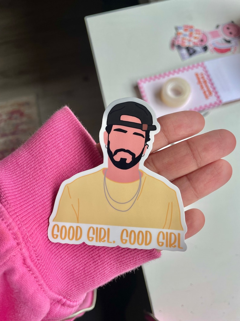 Puede incluir: Una pegatina con una ilustraci&oacute;n estilizada de un hombre con gorra de b&eacute;isbol negra y camisa amarilla. La pegatina tiene el texto "GOOD GIRL. GOOD GIRL" en letras naranjas.