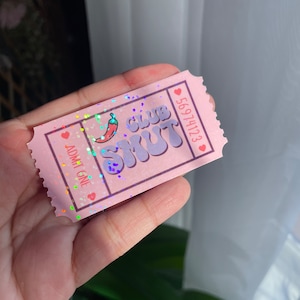 May include: A pink ticket with the text "CLUB SMUT" and a chili pepper graphic. The ticket also has the text "ADMIT ONE" and a serial number. The ticket is decorated with hearts and glitter.
