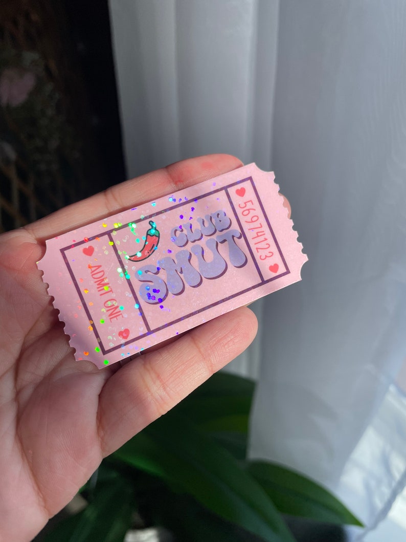 May include: A pink ticket with the text "CLUB SMUT" and a chili pepper graphic. The ticket also has the text "ADMIT ONE" and a serial number. The ticket is decorated with hearts and glitter.