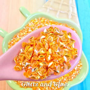 May include: Close-up of orange glitter in a pink spoon, with more glitter in a green dish. The dish is shaped like an ice cream cone. The text "Glitz and Glue" is visible.