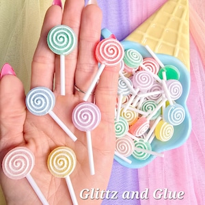May include: A collection of colorful, miniature lollipops with a spiral design. The lollipops are in various pastel colors, including green, pink, blue, and yellow, and are displayed in a blue dish and held in a hand. The text "Glitz and Glue" is visible.