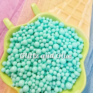 May include: A light green, heart-shaped dish filled with light blue, round beads. The dish is next to a yellow, waffle-patterned cone shape. The text "Glitz and Glue" is written on the dish.