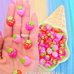 May include: A collection of strawberry-shaped embellishments in various shades of pink and red, with green leaf accents. Some are held in a hand, others in a light green ice cream cone-shaped dish. The text "Glitz and Glue" is visible.