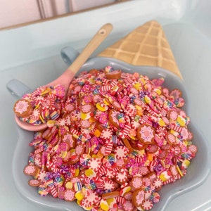 May include: A blue ceramic bowl filled with colorful candy-themed polymer clay sprinkles. The sprinkles are shaped like candy canes, donuts, and flowers. The bowl is sitting on a light blue tray with a waffle cone.