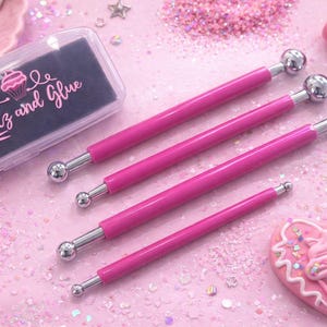 May include: A collection of pink craft tools, including four dotting tools with silver ball tips, a small rolling pin, and a heart-shaped decoration with a cupcake design. The tools are arranged on a pink surface with glitter and small beads. The text "Glitz and Glue" is visible.