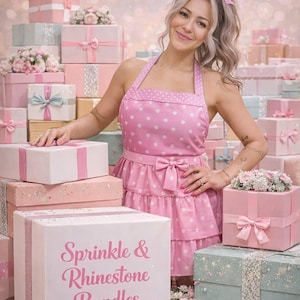 May include: A woman in a pink polka dot apron stands among a display of gift boxes. The boxes are in shades of pink, white, and teal, some with ribbons and floral arrangements. The front box reads "Sprinkle & Rhinestone Bundles."