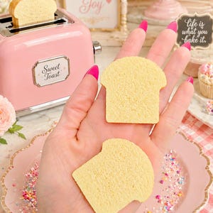 May include: A pink toaster with a slice of bread, and two bread-shaped sponges. The toaster has the words "Sweet Toast" on it. The image also includes a pink plate, sprinkles, and the text "Sprinkle a little joy" and "life is what you bake it."