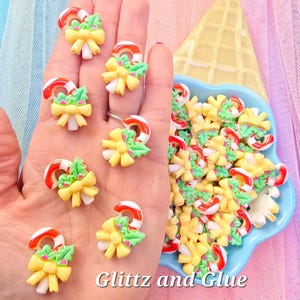 May include: A collection of Christmas-themed resin embellishments. The image shows candy cane and bow designs in red, white, yellow, and green. The text "Glitz and Glue" is visible in the image. The embellishments are in a blue dish and in a person's hand.