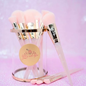 May include: A set of pink and gold makeup brushes in a clear glass container with a gold rim. The container has a sticker with the words "Glittz and Glue" and a cupcake graphic.