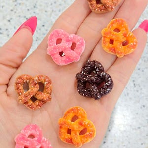 May include: A collection of miniature pretzel-shaped candies in various colors, including pink, orange, brown, and a speckled orange-brown. The candies are arranged on a light background, showcasing their detailed pretzel design.