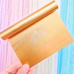 May include: A gold-colored pastry scraper with a rolled handle. The scraper has the text "Glitz and Glue" in a cursive font. The background is a soft gradient of pink, white, and blue.