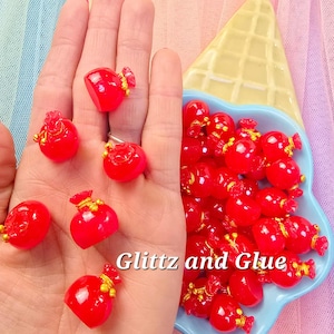 May include: A collection of small, glossy red resin charms shaped like pouches, some with gold-colored accents. The charms are displayed on a hand and in a blue dish, with the text "Glitz and Glue" visible. The background is a colorful, soft fabric.