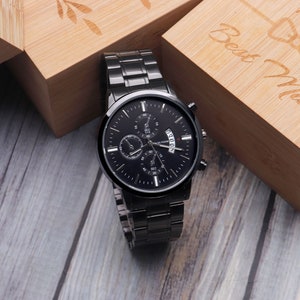 May include: A black chronograph wristwatch with a black face and silver hands. The watch is on a black metal band and is sitting on a wooden surface. The watch is in a box that says "Best Man" on it.