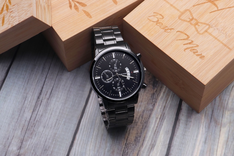 May include: A black chronograph wristwatch with a black face and silver hands. The watch is on a black metal band and is sitting on a wooden surface. The watch is in a box that says "Best Man" on it.
