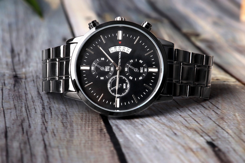 Puede incluir: Un reloj de pulsera cron&oacute;grafo negro con una correa de acero inoxidable. La esfera del reloj tiene tres subesferas y una ventana de fecha.