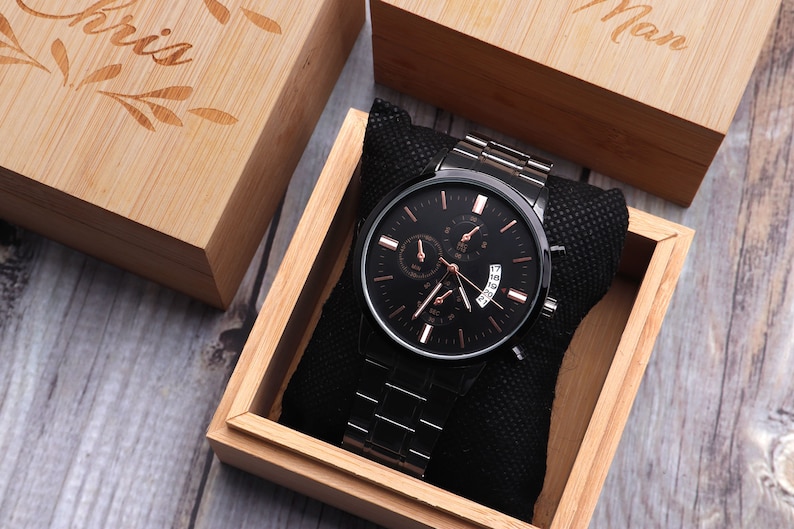May include: A black watch with a rose gold face and hands, in a wooden box with a black velvet lining. The box is engraved with the word "Man".