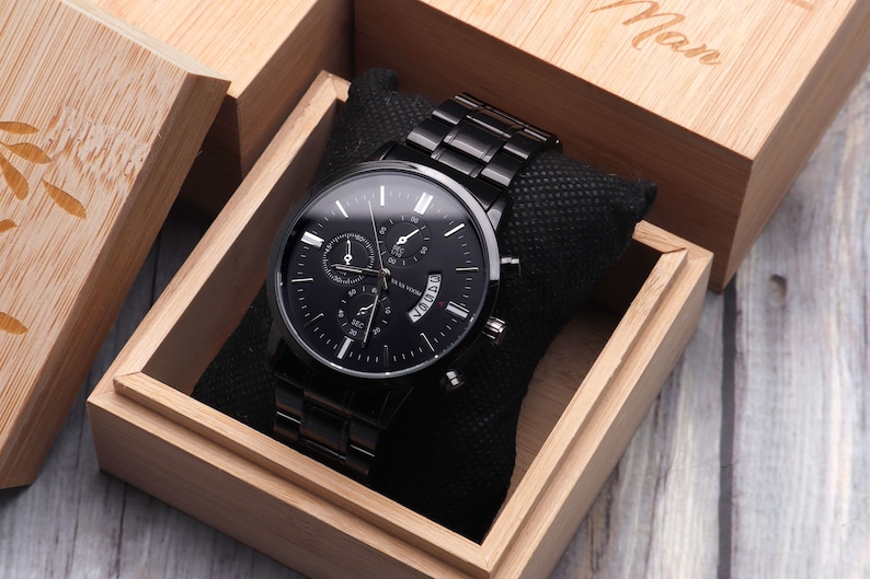 May include: A black watch with a black metal band in a wooden box. The watch has a black face with white markings and a date window. The box is made of light wood and has the word "Man" engraved on the top.