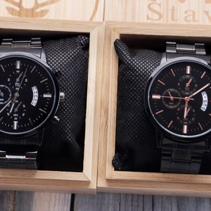 May include: Two black wristwatches with silver accents and black leather bands in wooden boxes. The watches have multiple chronograph dials and a date window.