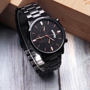 Puede incluir: Un reloj de pulsera negro con una esfera y agujas de oro rosa. El reloj tiene una correa de metal negro y una ventana de fecha.