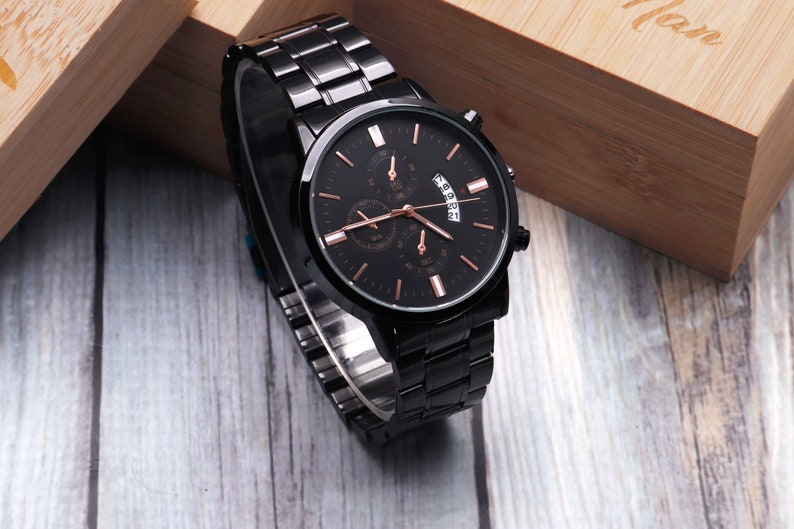 Puede incluir: Un reloj de pulsera negro con una esfera y agujas de oro rosa. El reloj tiene una correa de metal negro y una ventana de fecha.