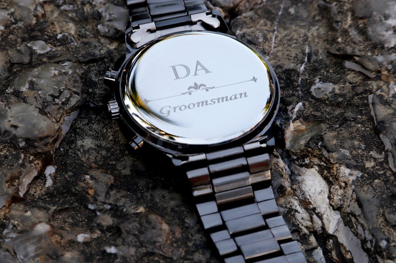 Puede incluir: Reloj de metal negro con una parte posterior plateada grabada con las iniciales "DA" y la palabra "Groomsman".