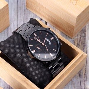 Puede incluir: Un reloj negro con esfera y agujas de oro rosa, una ventana de fecha y una correa de metal negro. El reloj est&aacute; en una caja de madera.