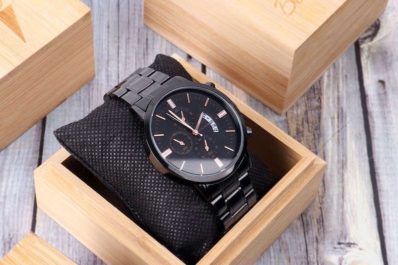Puede incluir: Un reloj negro con esfera y agujas de oro rosa, una ventana de fecha y una correa de metal negro. El reloj est&aacute; en una caja de madera.