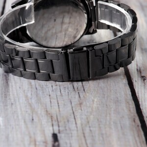 May include: A black metal watch band with a clasp closure. The band is made of linked metal pieces.