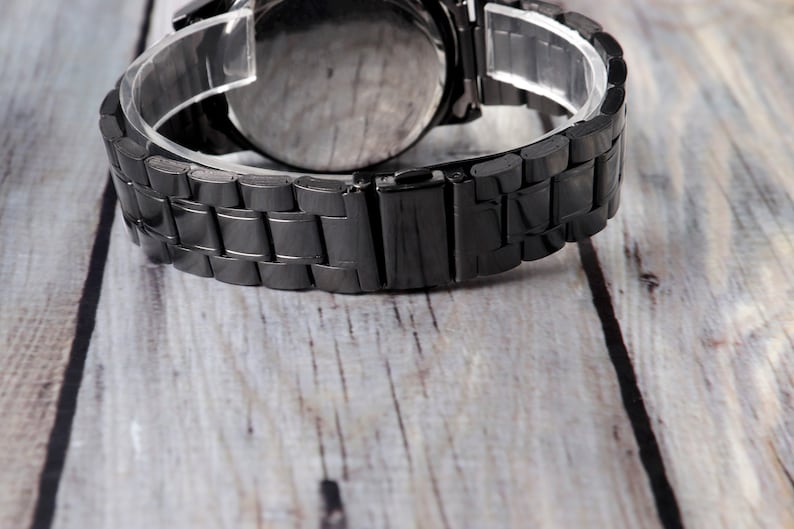 May include: A black metal watch band with a clasp closure. The band is made of linked metal pieces.
