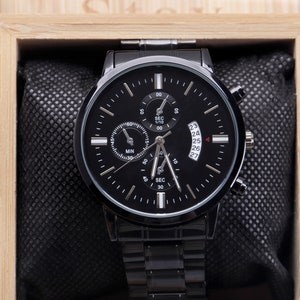 May include: A black wristwatch with a black face and silver hands. The watch has a date window and a chronograph function. The watch is in a wooden box.