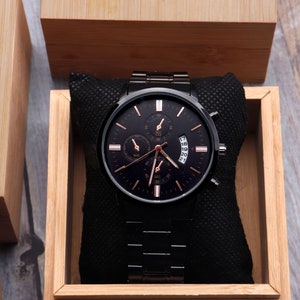 May include: A black chronograph wristwatch with rose gold accents and a black leather band, displayed in a wooden box.