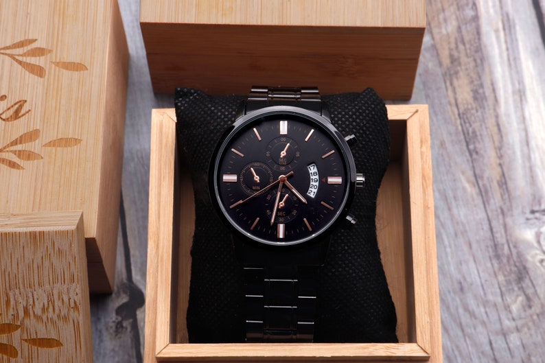 May include: A black chronograph wristwatch with rose gold accents and a black leather band, displayed in a wooden box.