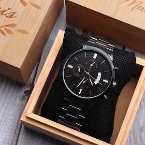 Puede incluir: Un reloj negro con esfera y agujas de oro rosa, en una caja de madera con forro de terciopelo negro. La caja est&aacute; grabada con la palabra "Hombre".