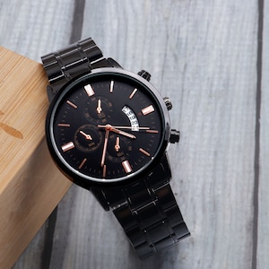 May include: A black wristwatch with a rose gold face and hands. The watch has a black metal band and a date window.