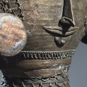 May include: A bronze sculpture of a face with intricate details. The face has a large nose and a small mouth. The sculpture is decorated with a series of bands of raised and recessed lines.